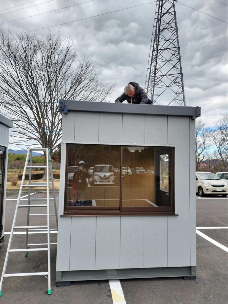 70歳を迎える社員がプレハブの屋根で測定作業を行っている様子。